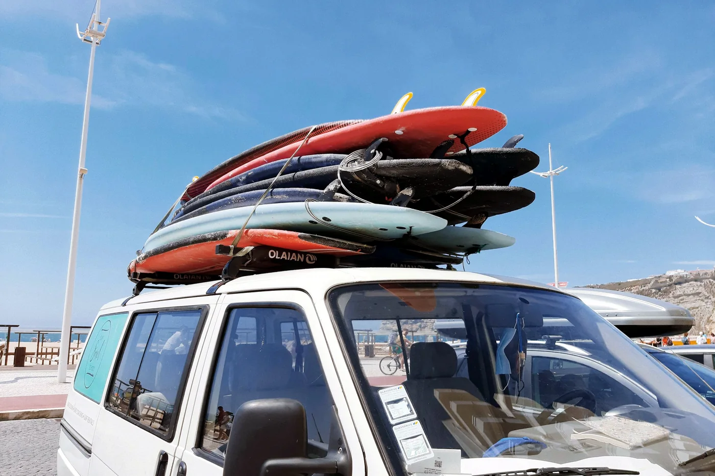 Стопка сёрфбордов закреплена на крыше светлого фургона, рядом море