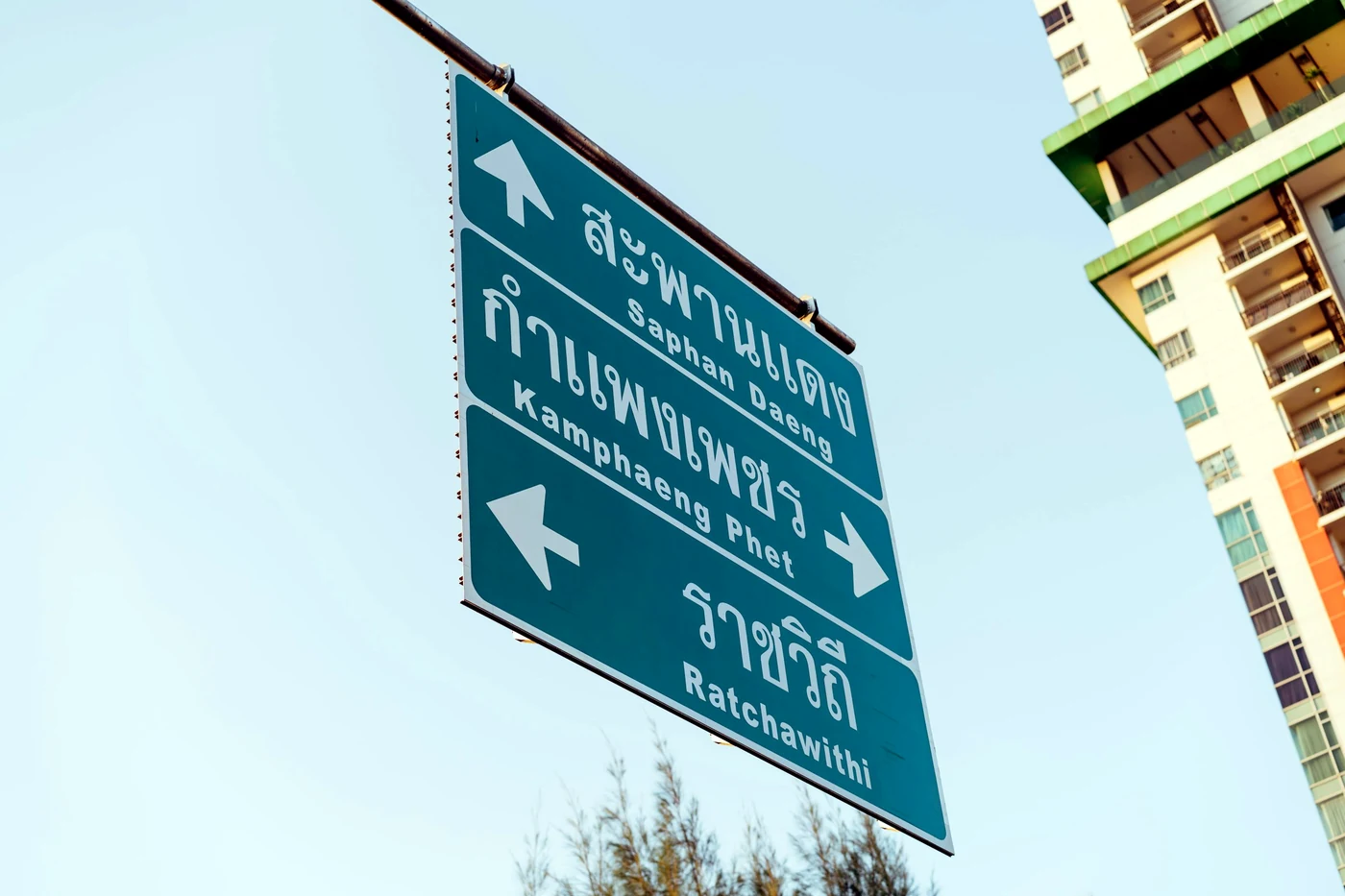 Зелёный дорожный указатель с надписями на тайском и латинице и стрелками на фоне неба